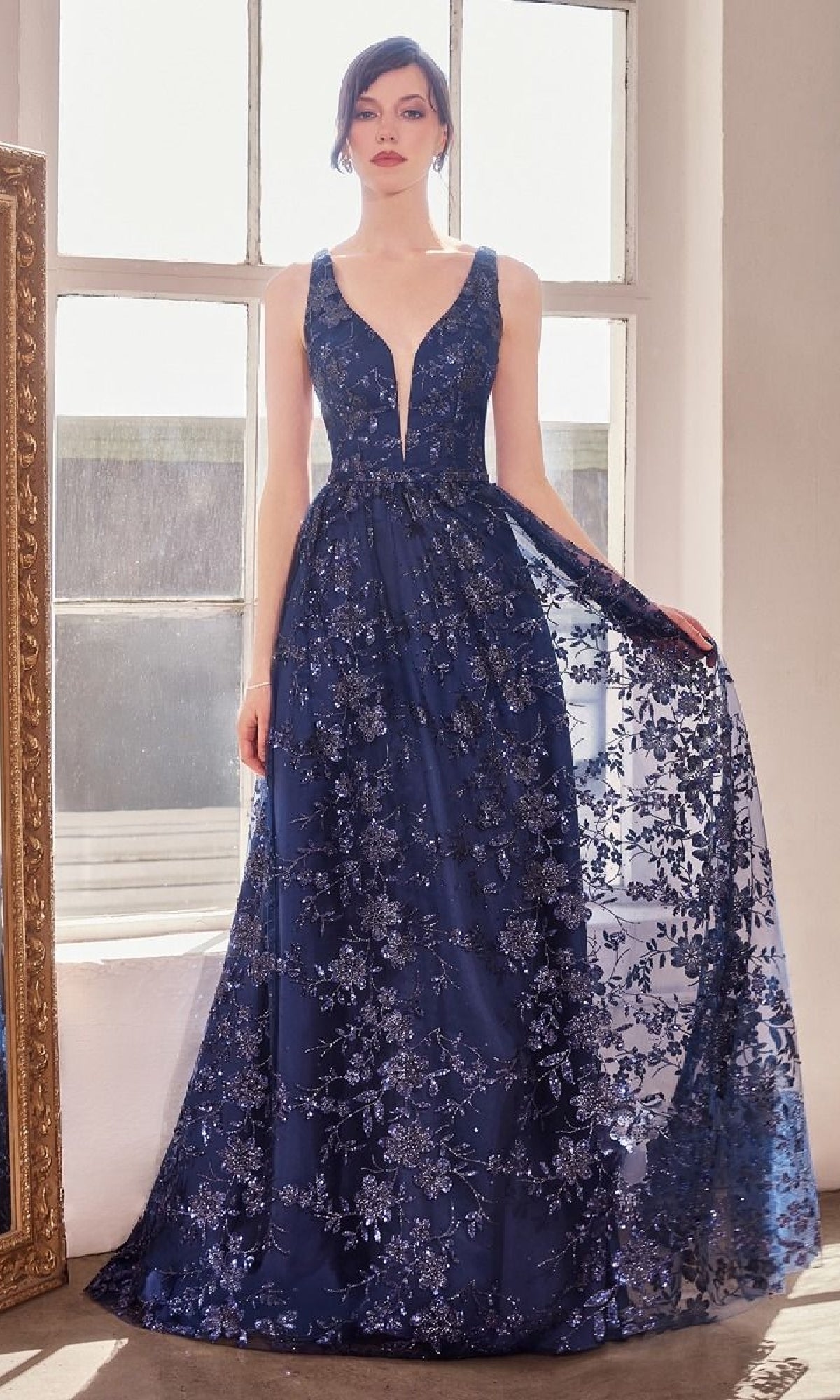 A woman stands in front of large windows wearing the Amoris T606 Embroidered-Lace Long Prom Dress, a sleeveless, deep V-neck navy gown with floral lace and sequin embellishments.